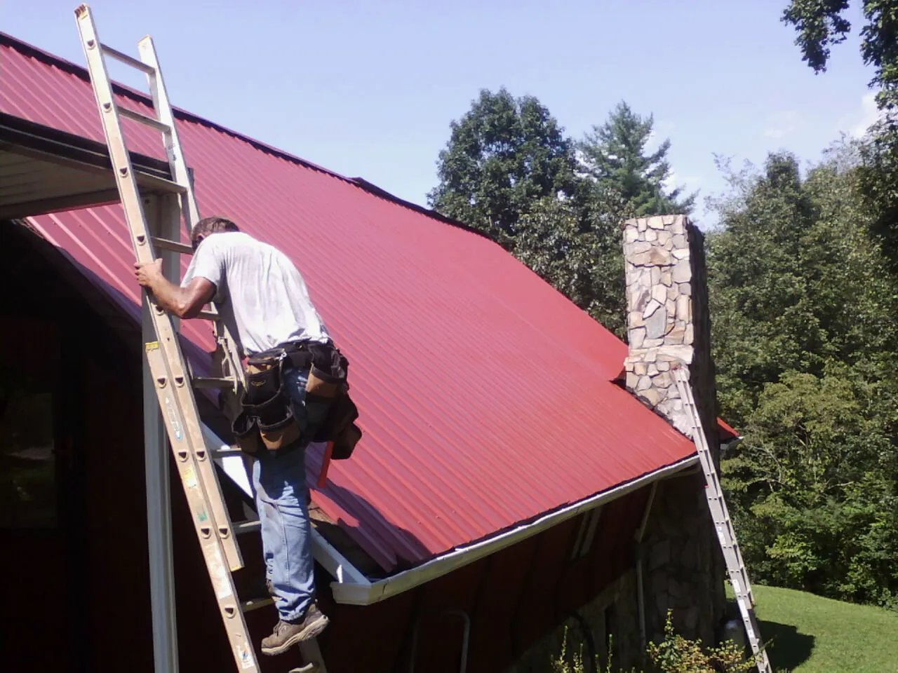 Roofer on a ladder beside a red metal roof and stone chimney — Soffit & Fascia Repair by Roofing Brothers in Syracuse